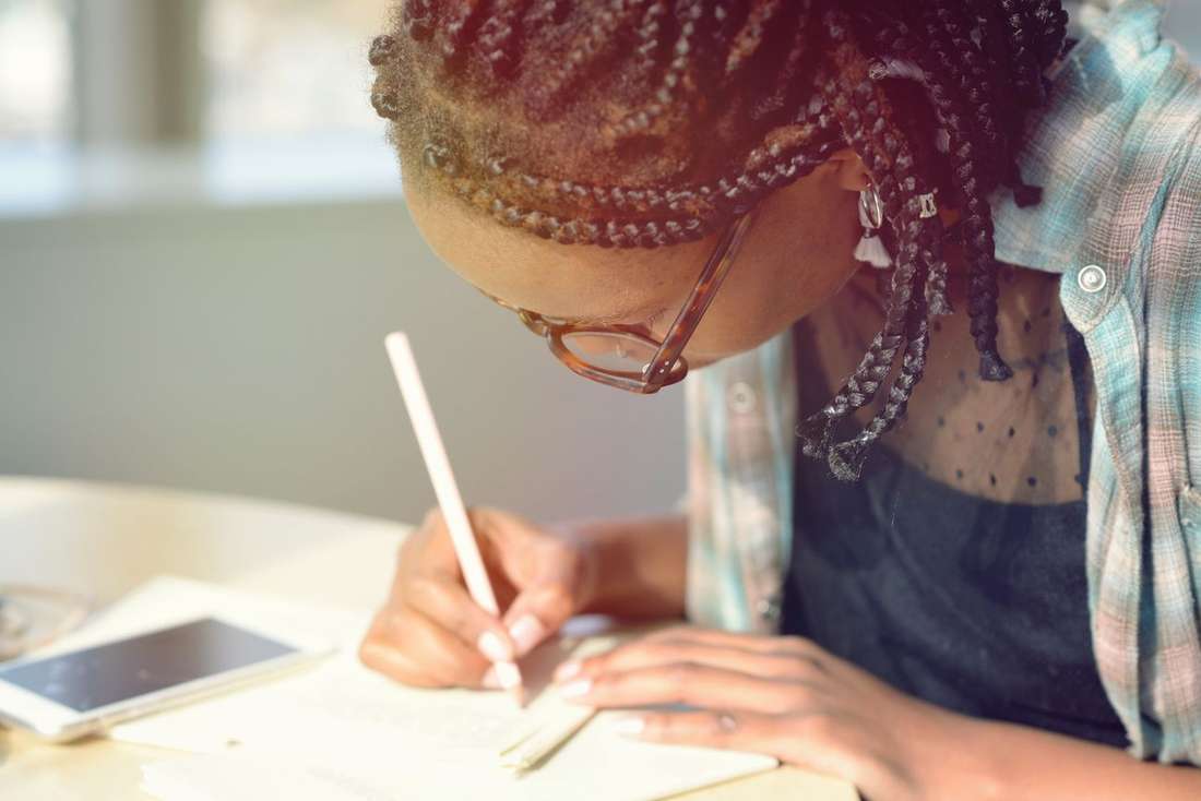 A young Black woman with braids and glasses writing intently with a pen
