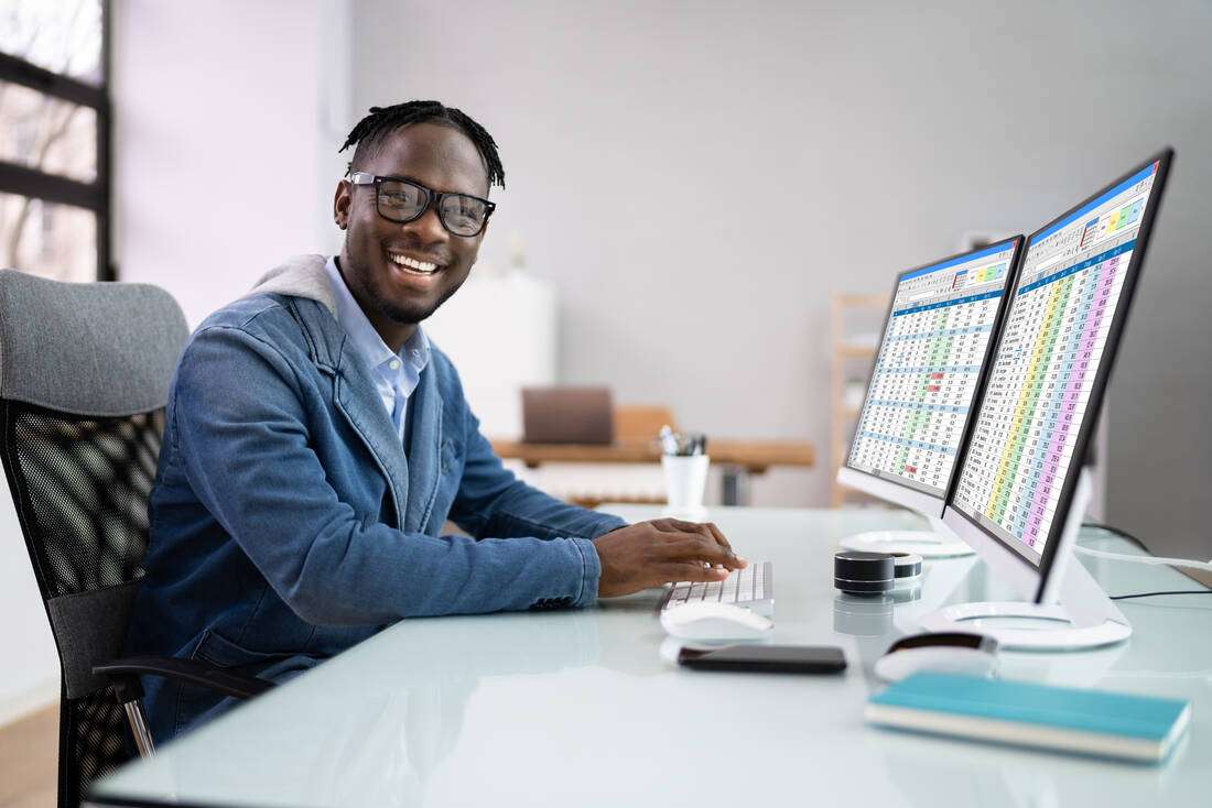 A smiling man in glasses working at a computer with two screens