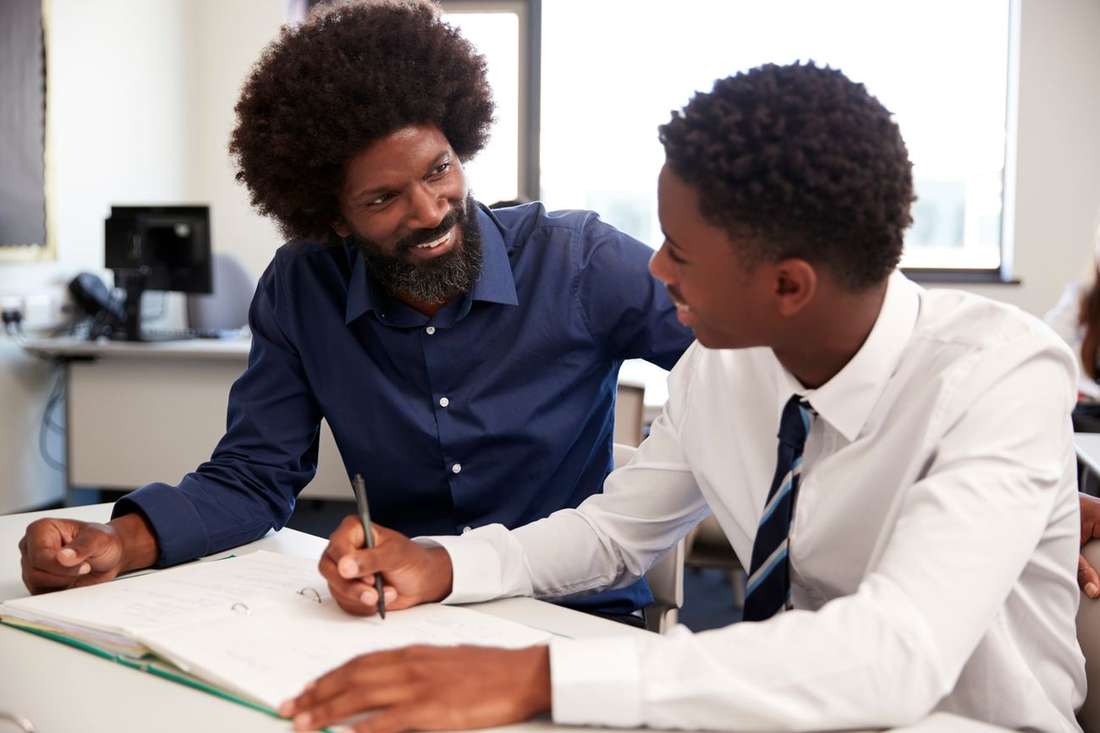 A Black mentor and a young man in a shirt and tie talking together over his written work
