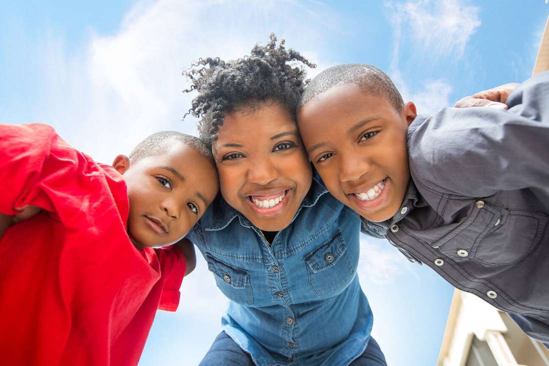 A mother hugging two smiling children, seen from below against a blue sky
