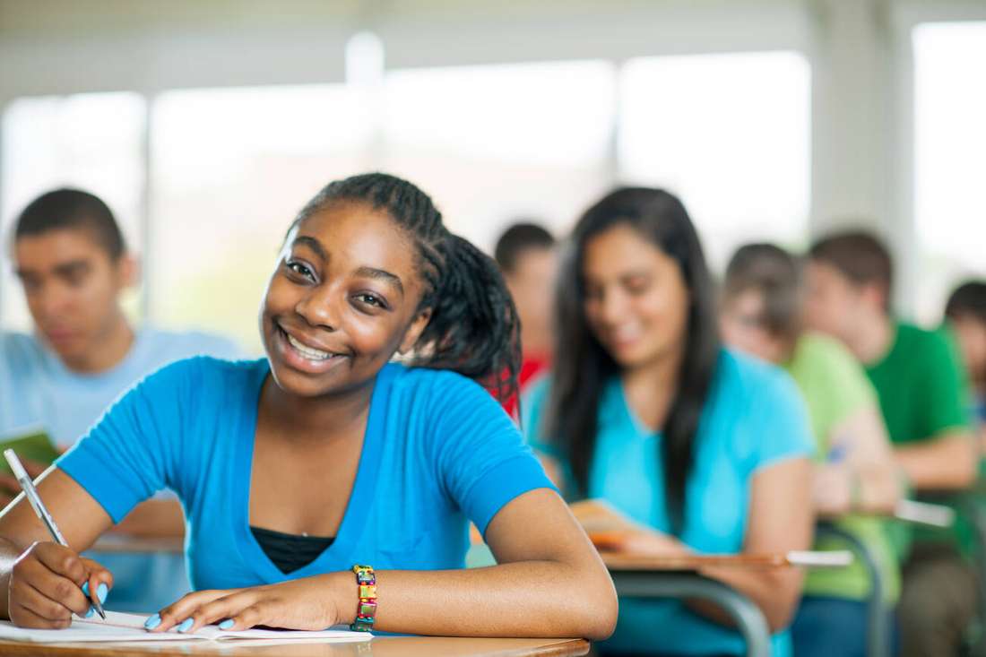 A smiling learner writing at her desk in a busy classroom