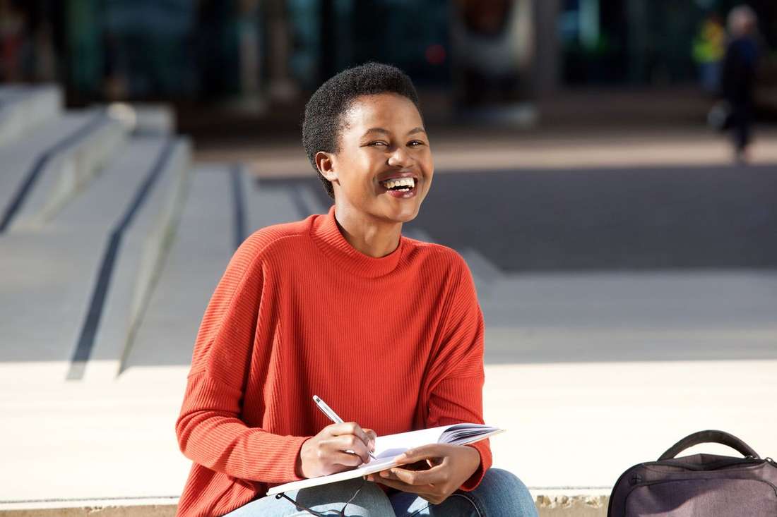 A joyful young Black woman in a red sweater laughing as she writes in her notebook on sunny steps
