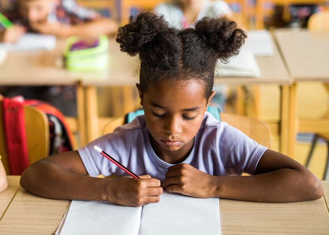 A young Black girl with her hair in puffs, writing carefully in her notebook at a school desk