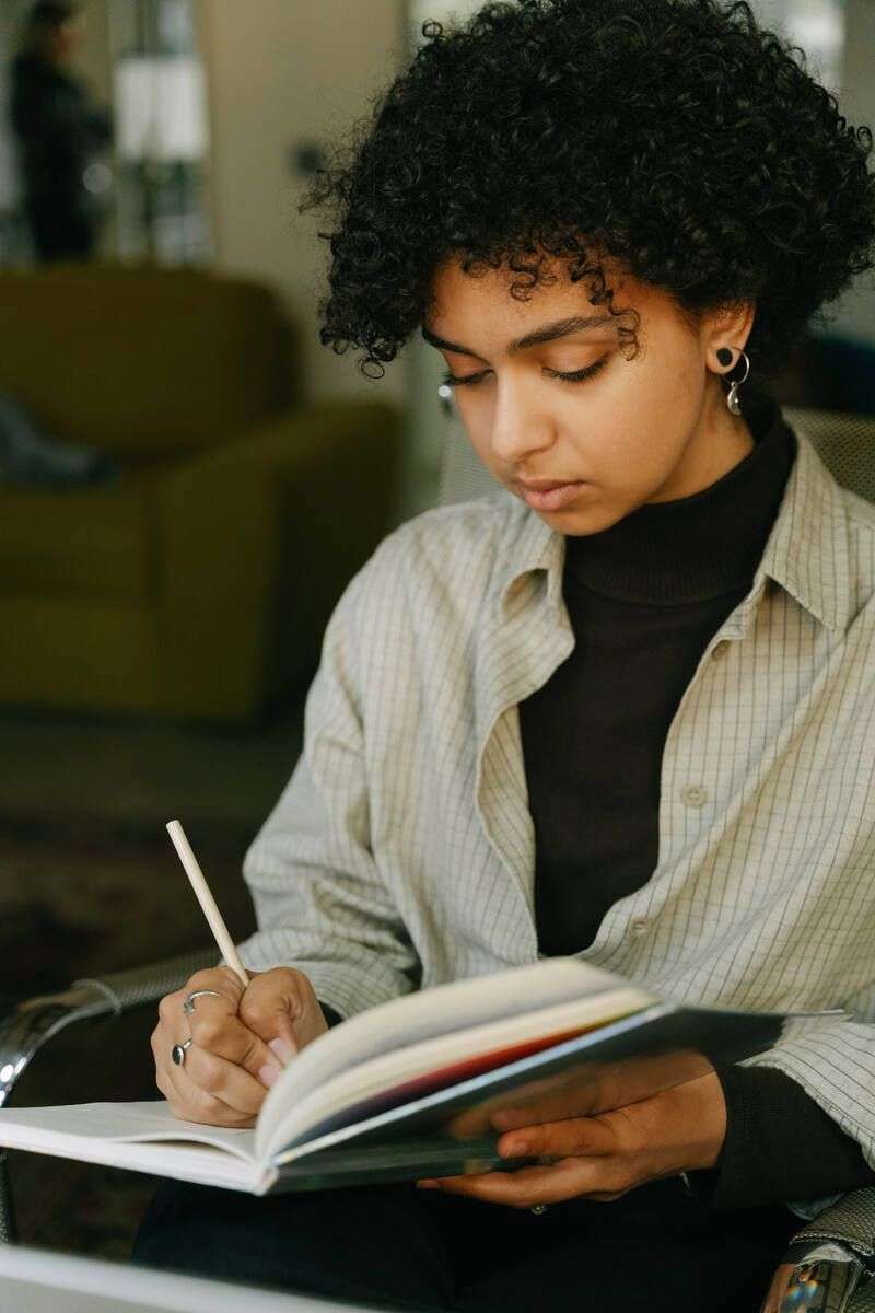 A young Black woman with curly hair writing thoughtfully in her notebook